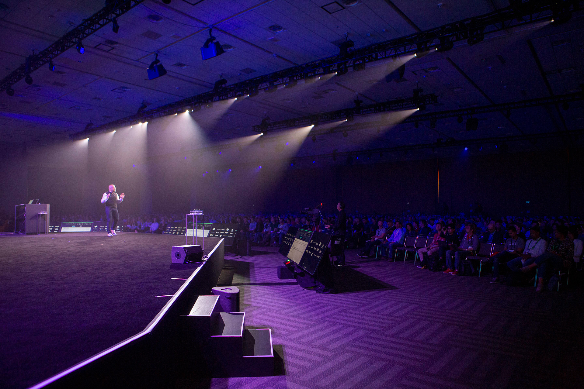 Wide shot of convention center ballroom with speaker under dramatic spotlights and seated audience